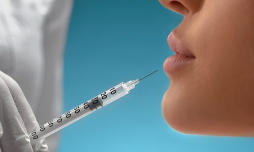 Side view of cosmetic botox injection in female lips. Close up of cosmetologist using small syringe with special liquid to make lips bigger, isolated on blue. Concept of cosmetology, beauty.